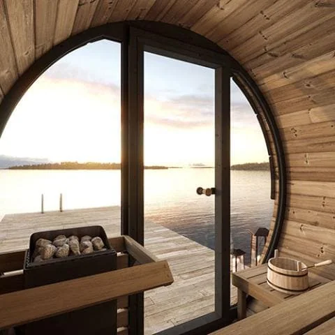 Interior view of barrel sauna with glass door overlooking lake sunset, featuring sauna rocks, wooden bench, and traditional water bucket