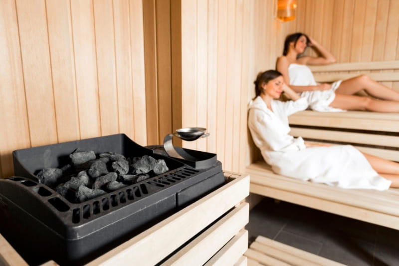 Modern electric sauna heater with hot stones in foreground, two women relaxing on wooden benches in background