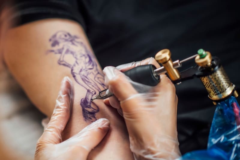 A female tattoo artist carefully tattooing a detailed design on a client's arm.