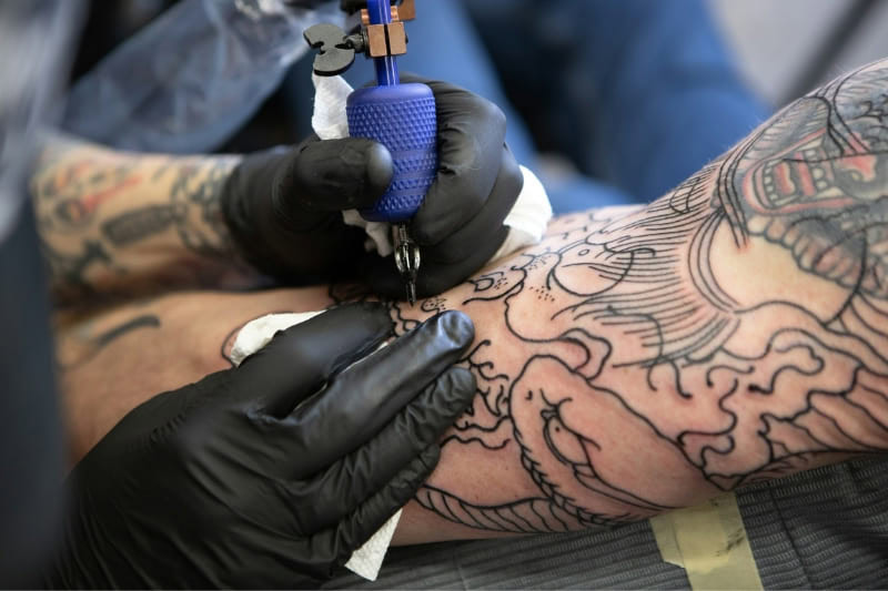 Close-up of a tattoo artist adding intricate linework to an arm tattoo, with a tattoo machine in use.