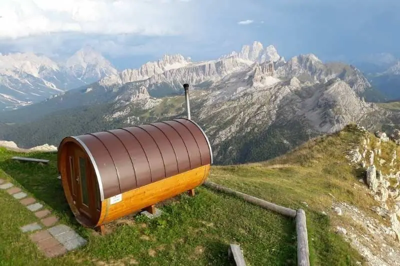 Panoramic mountain view barrel sauna with metal roof and wooden base, set against dramatic Alpine landscape