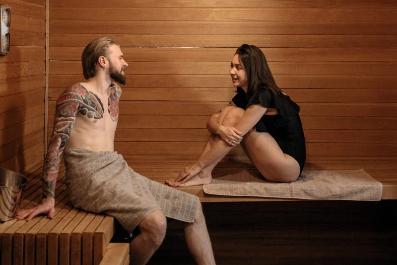 A man with an extensive chest and arm tattoo sitting in a sauna, engaging in conversation with a woman.