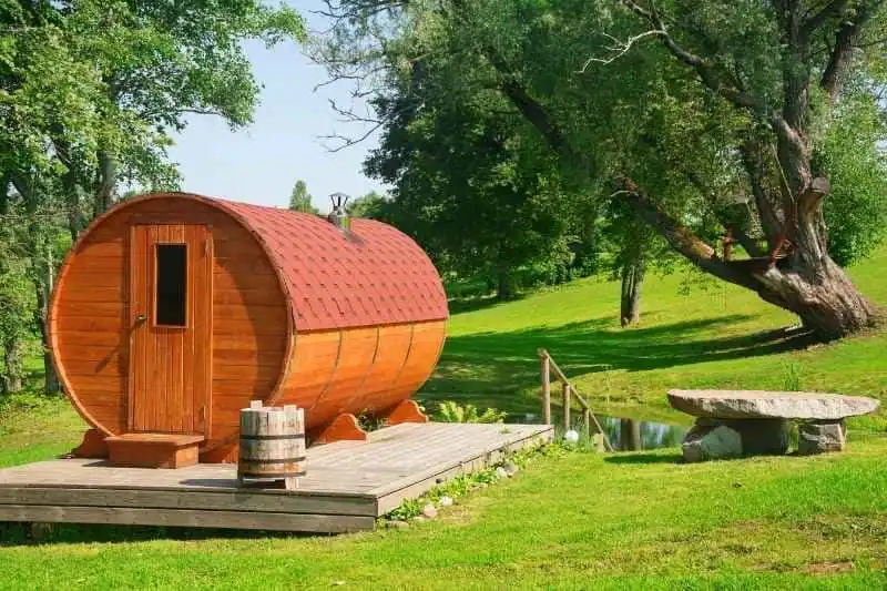 Red cedar barrel sauna in garden setting with wooden deck platform and natural landscaping, perfect for backyard sauna construction