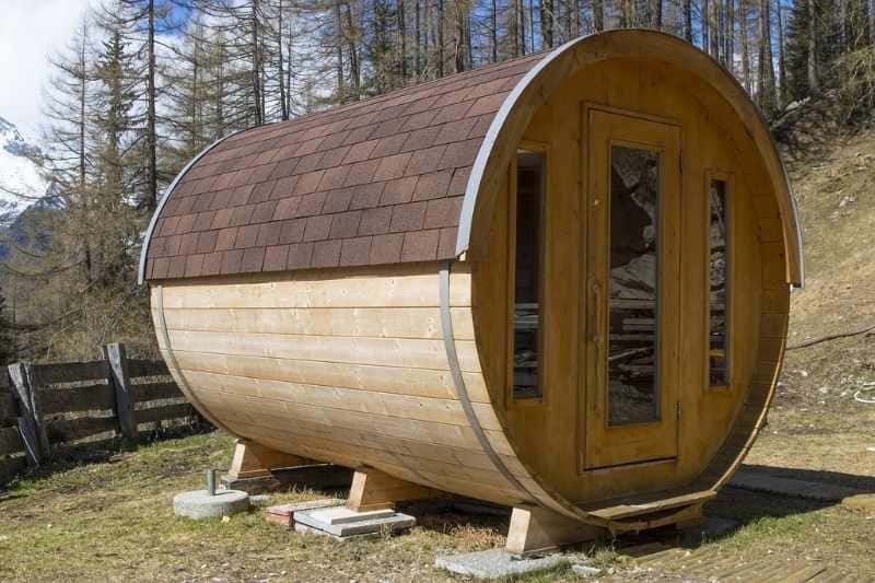 Wooden barrel sauna with shingled roof installed on hillside among pine trees, featuring glass door and natural wood finish