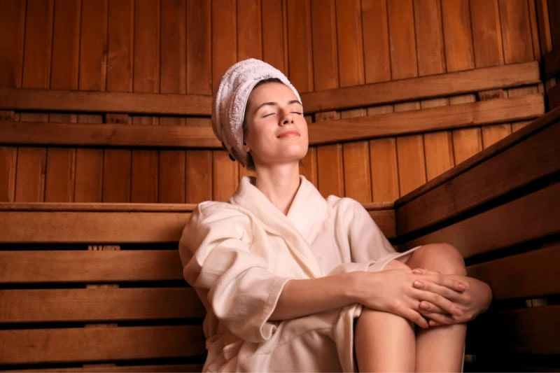 Woman wearing white robe and hair towel relaxing on wooden bench in traditional sauna