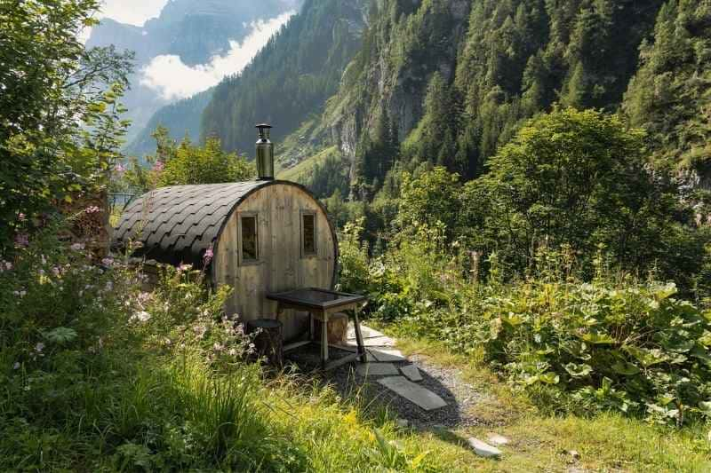 Wooden barrel sauna with shingle roof nestled in lush mountain valley, featuring natural garden path and scenic alpine backdrop