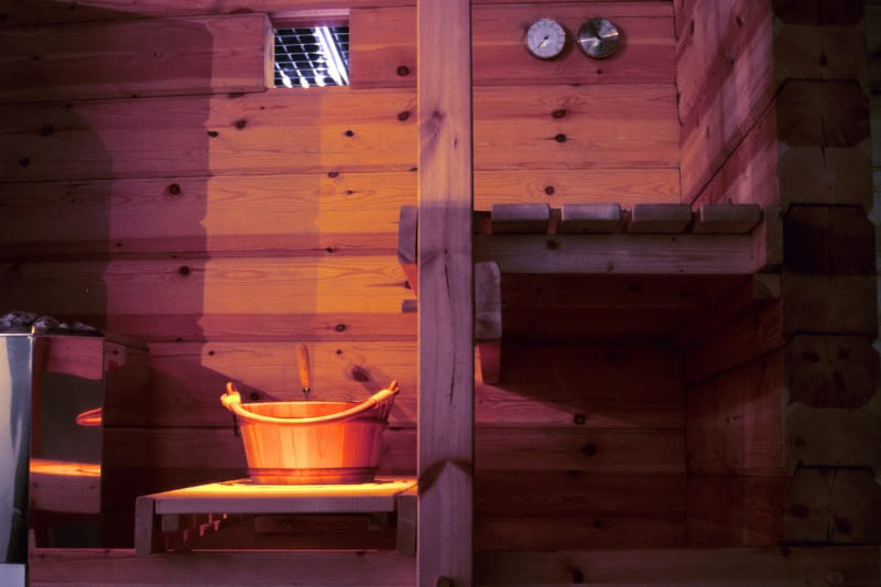 Traditional wooden sauna interior with water bucket and wall-mounted temperature gauges, illuminated by warm ambient lighting