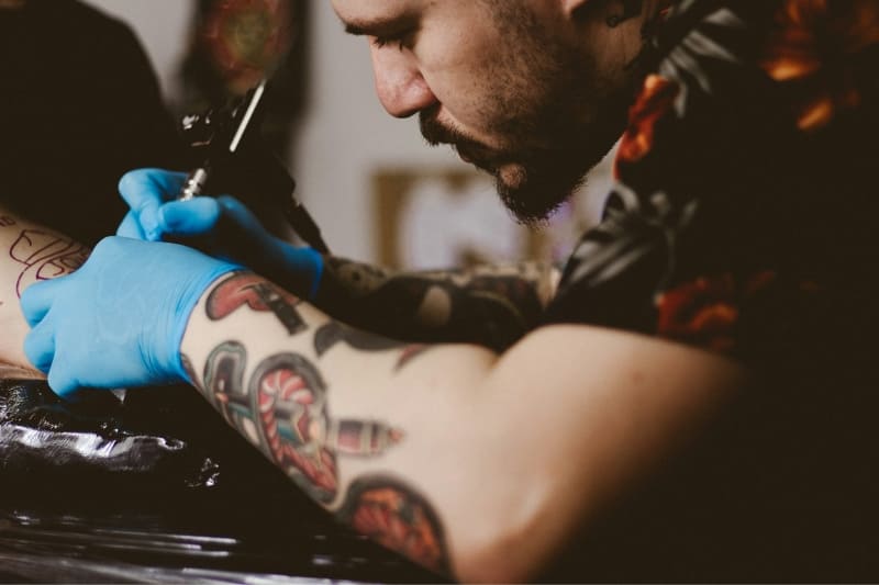 A tattoo artist focused on creating a detailed arm sleeve tattoo, wearing gloves for hygiene.