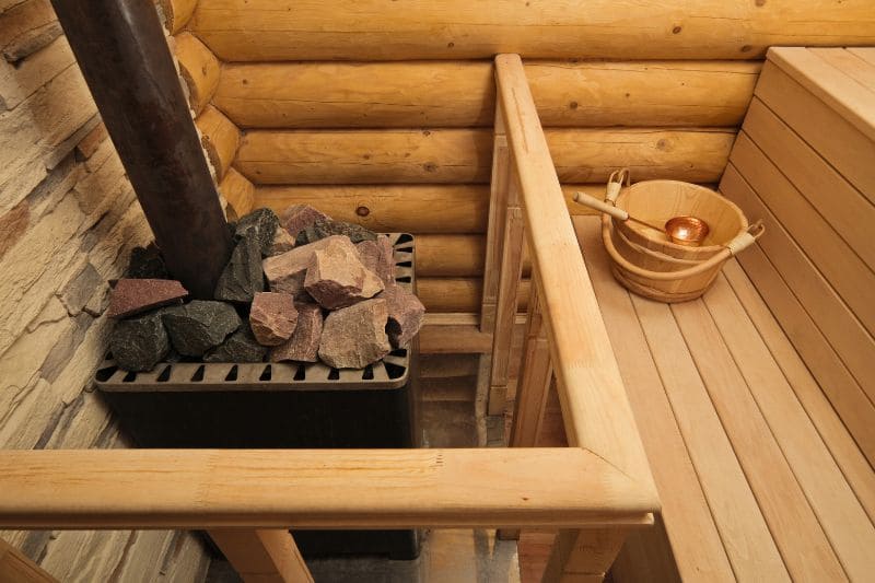 Traditional wood-burning sauna stove with heating stones and wooden water bucket in rustic log cabin sauna