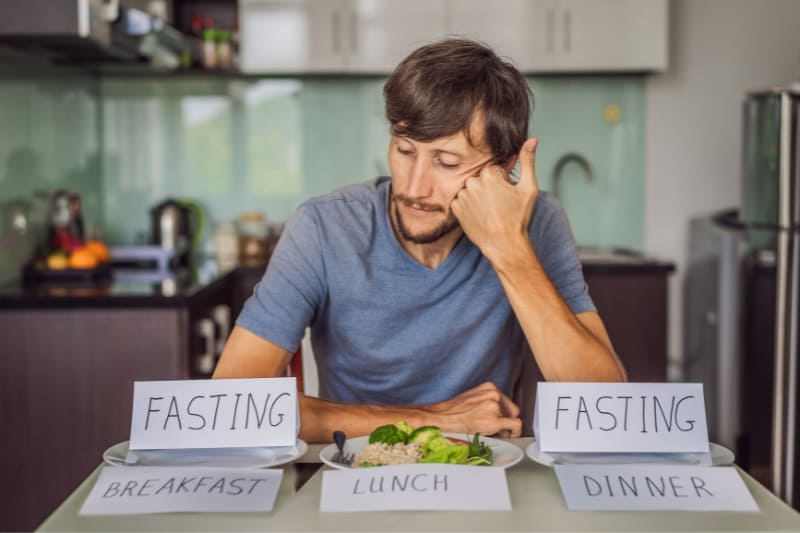 Man looking tired at meal planning table with cards labeled 'Fasting' for breakfast, lunch, and dinner, demonstrating intermittent fasting schedule