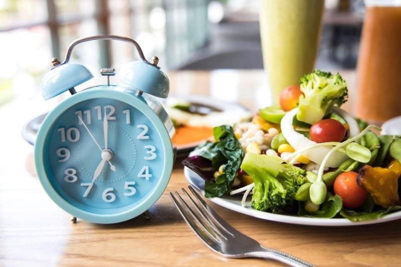 Blue alarm clock at meal time next to fresh healthy salad plate, showing intermittent fasting schedule