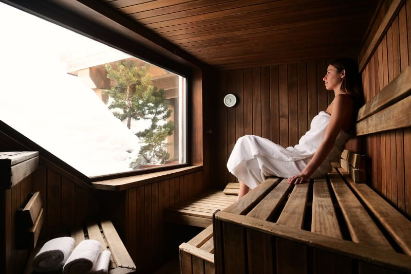 Woman relaxing in wooden sauna with large window, wearing white towel, demonstrating stress-reduction benefits of sauna therapy