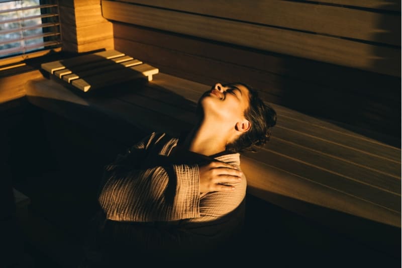 Woman lying back on sauna bench with relaxed expression, bathed in warm golden light streaming through wooden slats