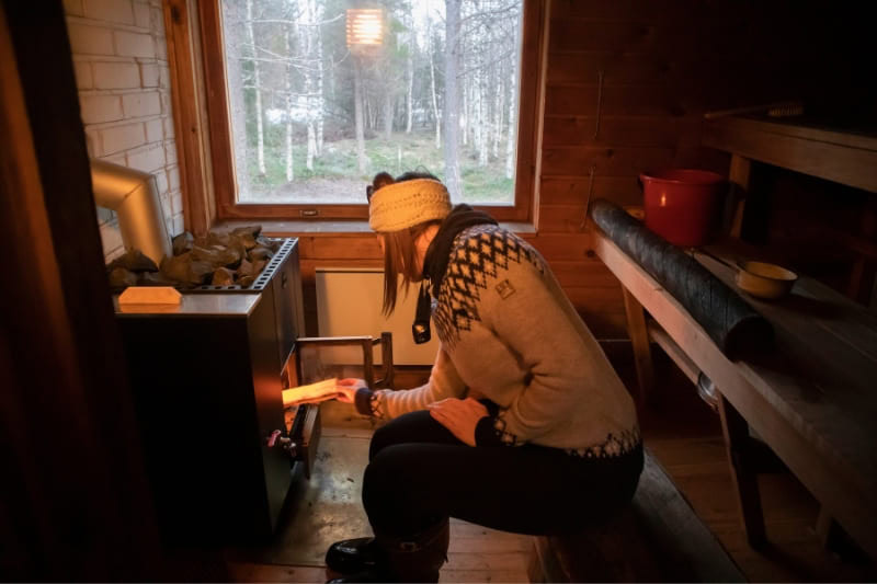 Woman tending to wood-burning sauna stove in cozy cabin setting with winter forest view through window