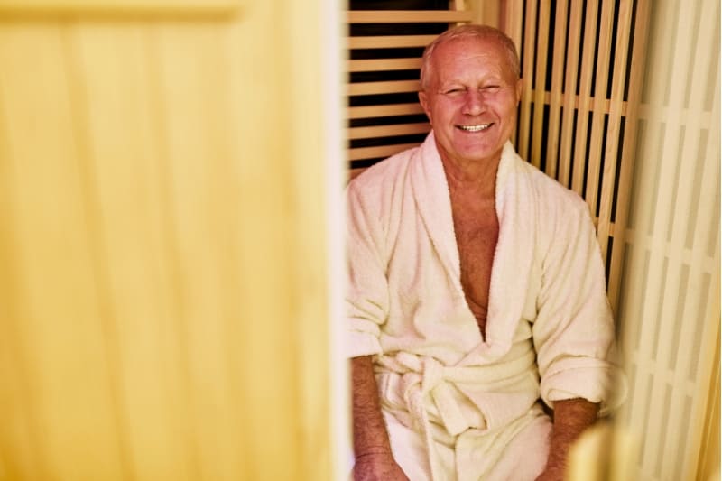 Older man in white bathrobe smiling warmly while enjoying an infrared sauna