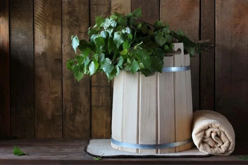 Traditional wooden sauna bucket with fresh birch branches and rolled towel against rustic wood paneling of the sauna