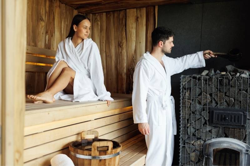 A couple in white robes inside a sauna with a traditional sauna heater, with the lady sitting on the wooden bench and the man pouring water on the sauna rocks