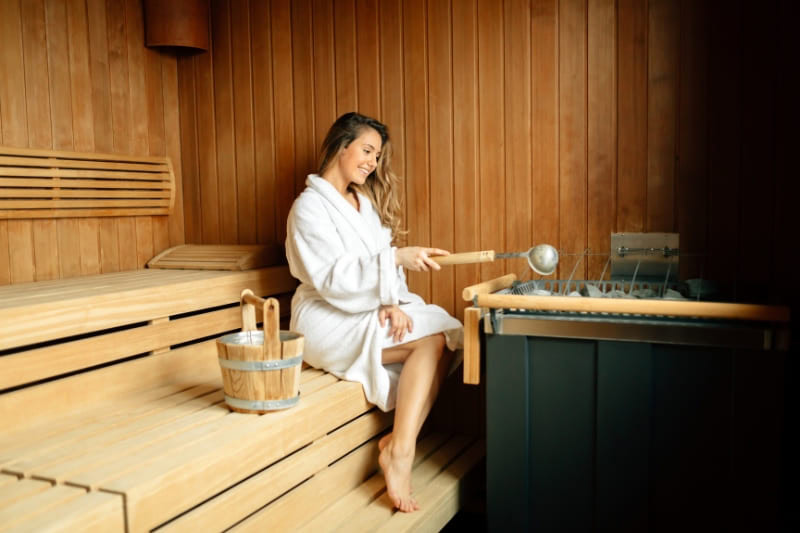 Woman in white robe relaxing on wooden bench in traditional Finnish sauna with water bucket and ladle