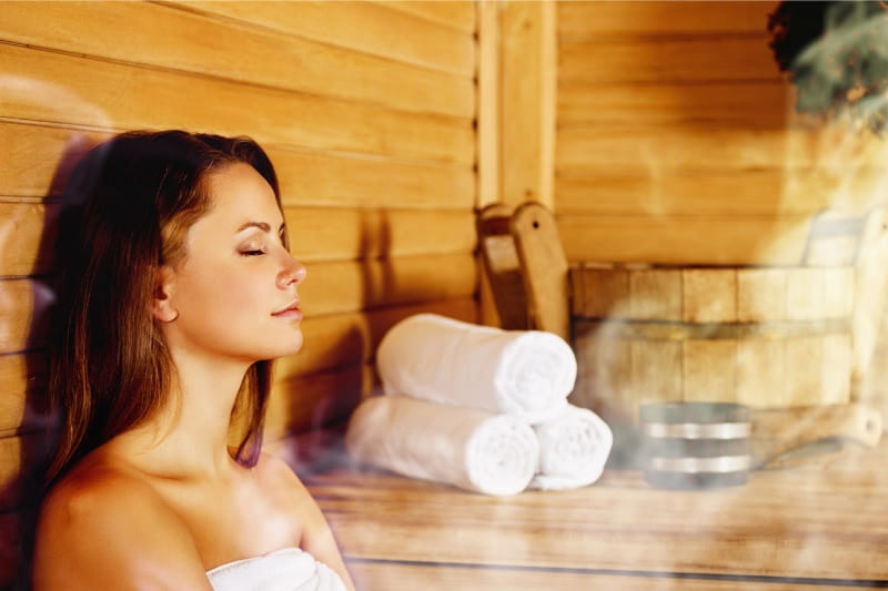 Woman relaxing in wooden sauna wrapped in white towel with rolled towels in background