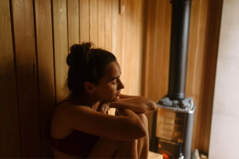 Woman relaxing in wooden sauna, sitting against wall near wood-burning stove.