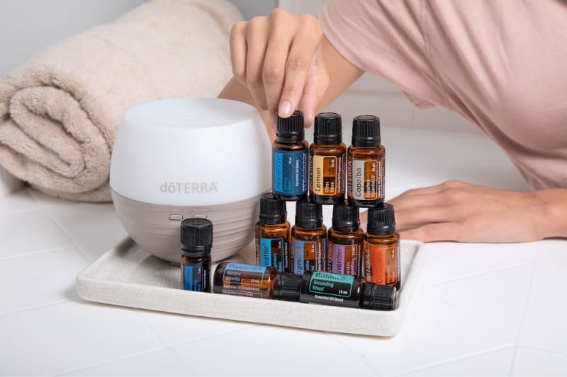 Modern white essential oil diffuser with collection of doTERRA oils arranged on ceramic tray, alongside plush towels in spa setting