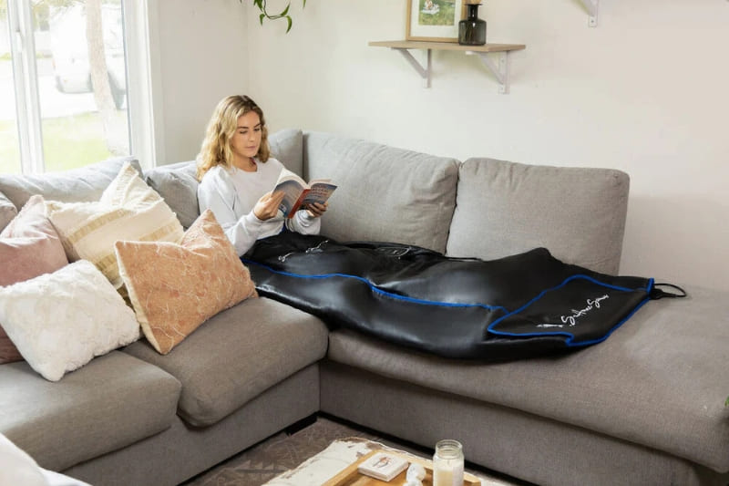 Woman relaxing on grey couch with book while using infrared sauna blanket in home setting