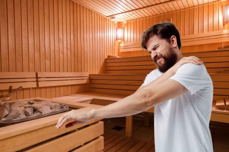 A man grimaces while rubbing his shoulder in a sauna, appearing to be experiencing muscular pain or soreness that the sauna heat may help alleviate.