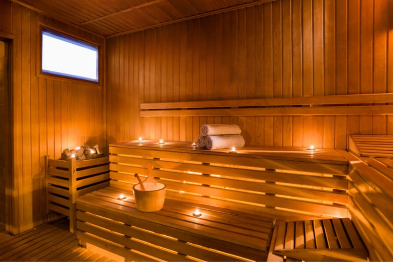 Modern fitness facility sauna with natural light window, atmospheric candles, wooden bucket, and stacked towels for guest comfort