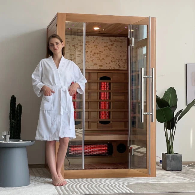 woman in white bathrobe standing next to a compact infrared sauna cabin with glass door and red heating elements in home setting