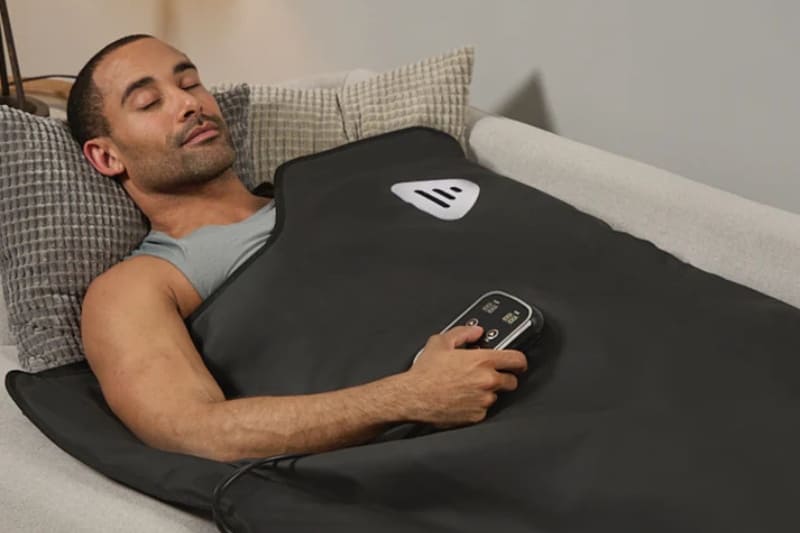 Man using an infrared sauna blanket while resting on couch with wired remote control in hand