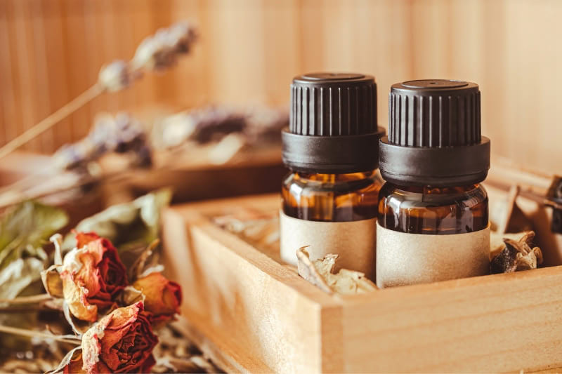 Two amber essential oil bottles in wooden box with dried orange roses and botanical elements, captured in warm lighting against wooden background