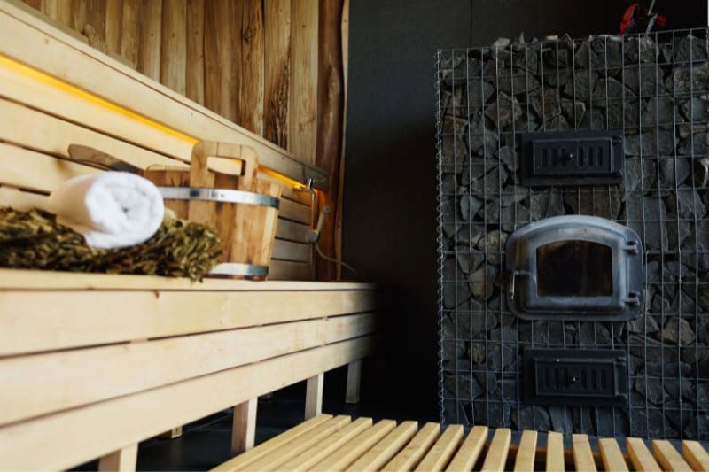 A metal wood fired sauna stove filled with rocks sits inside a wooden sauna, next to tiered benches.