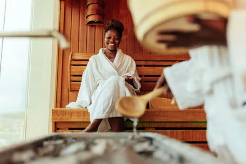 Woman in white bathrobe smiling inside a wooden traditional sauna, enjoying relaxation time