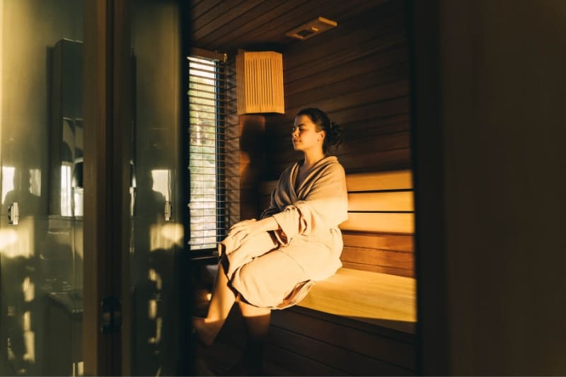 A woman wearing a muslin robe sits alone on the wooden bench inside a sauna, appearing relaxed.