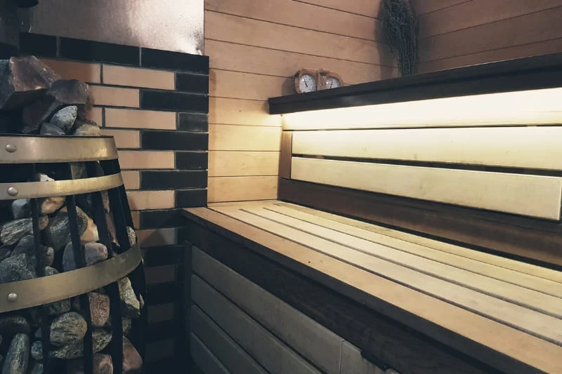 Contemporary sauna design with wooden benches, decorative brick wall, stone-filled heater, and ambient lighting for relaxation.