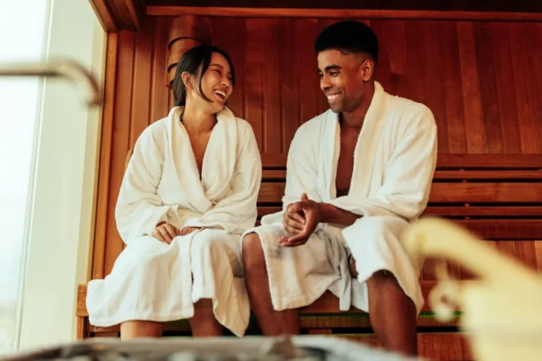 Happy couple in white bathrobes enjoying the health benefits of sauna therapy, smiling and conversing in a wooden sauna room.