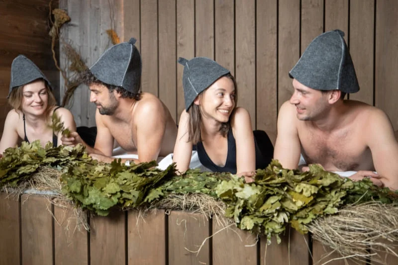 A group of friends relaxing in a traditional wooden sauna wearing gray felt sauna hats, demonstrating the social aspect of sauna culture