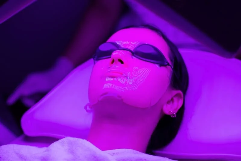 Woman receiving purple LED light therapy facial treatment with protective eyewear and hydrating mask for skin rejuvenation