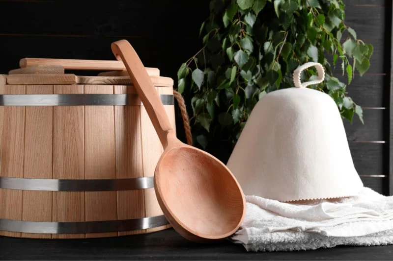 Essential sauna accessories including a wooden bucket, ladle, felt hat and birch leaves - traditional equipment for therapeutic sauna sessions.