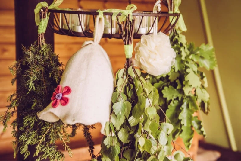 Sauna accessories hanging from a wooden rack, including a white felt sauna hat, birch branches (vihta), and bath accessories for authentic Finnish sauna experience