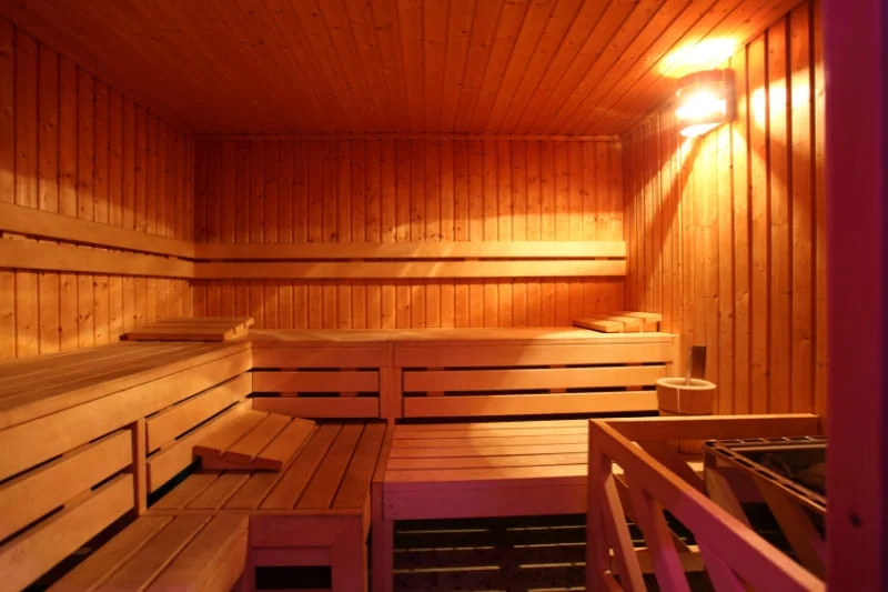 Interior of a traditional Finnish wooden sauna with tiered benches and warm lighting, showing empty seating areas and sauna heater.