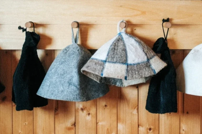 Various styles of wool and felt sauna hats hanging on wooden wall hooks, including solid gray, checkered pattern and black options for head protection