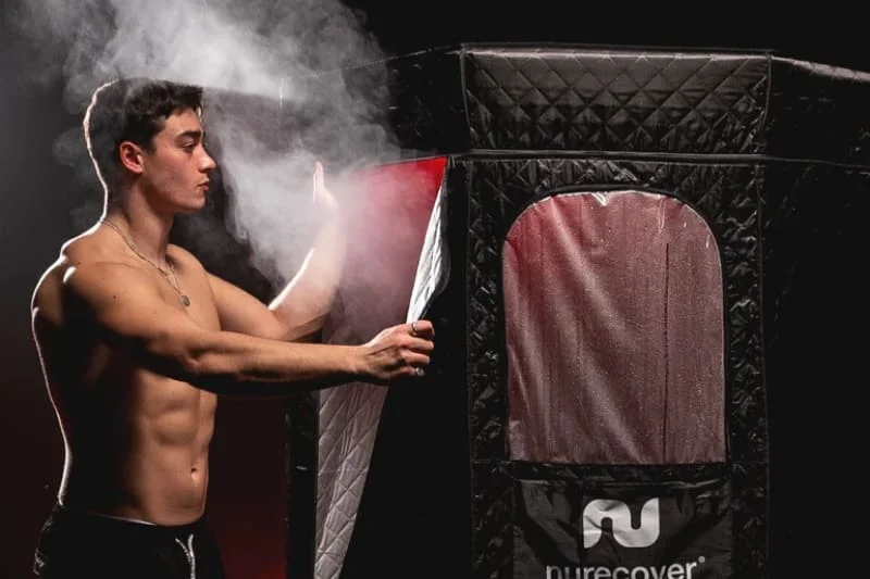 Shirtless man opening the black quilted Nurecover sauna with visible steam escaping from the opening