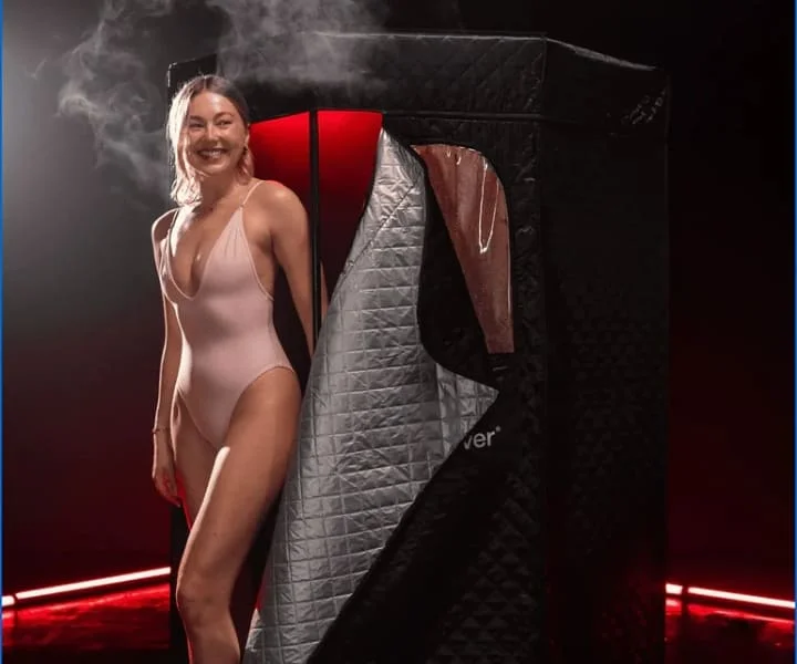 Woman in pink swimsuit standing beside open Nurecover sauna with visible steam and red interior lighting