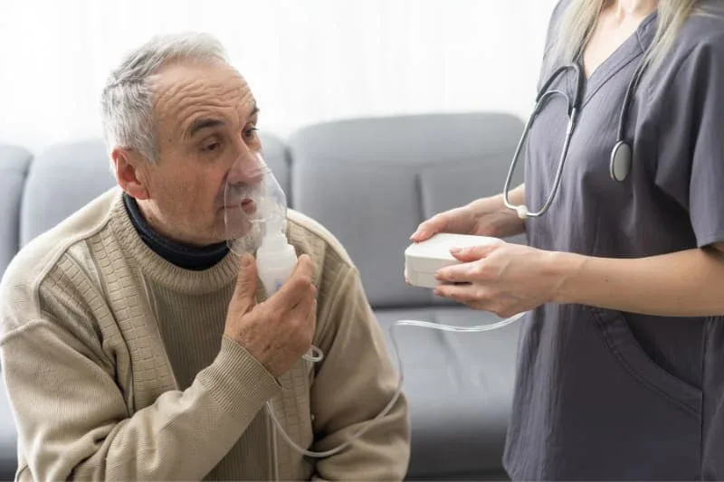 Elderly patient using a nebulizer for COPD treatment with medical professional assistance.