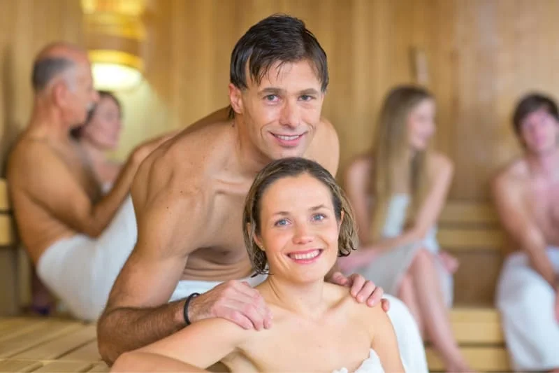 Young couple enjoying a traditional sauna experience with other sauna-goers in the background, demonstrating the social aspect of family sauna culture with everyone using towels for modesty.