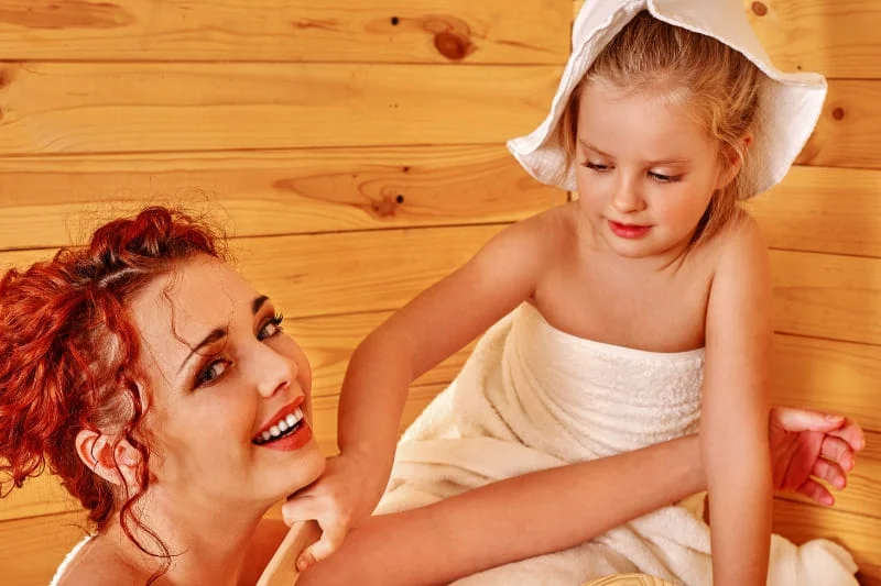 Mother and young daughter sharing an intimate bonding moment in a traditional pine sauna, with the child wearing a protective towel hat and both wrapped in white towels, showing proper sauna safety for kids.