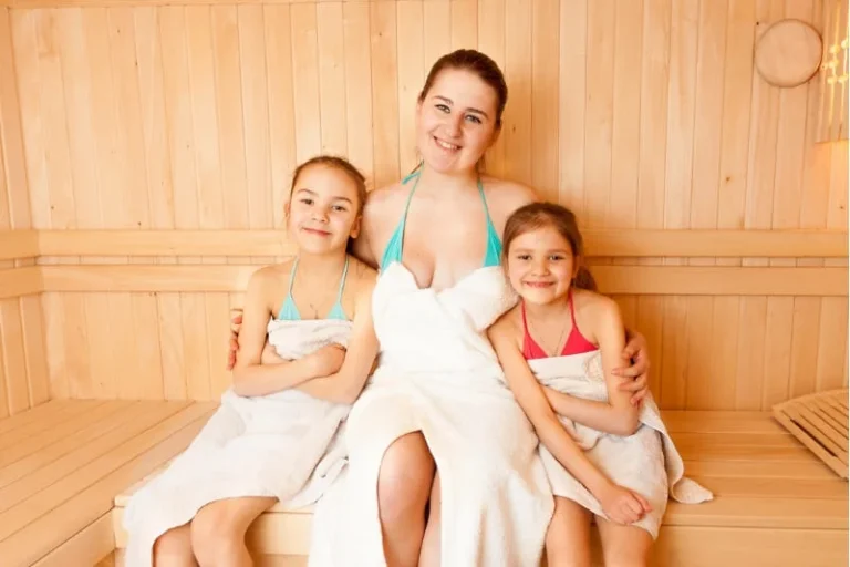 Mother and two daughters enjoying quality family time in a wooden sauna, all smiling and wrapped in white towels, demonstrating age-appropriate sauna use for children in a safe environment.