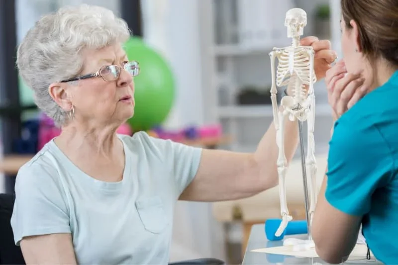 Senior woman with glasses discussing osteoporosis with a healthcare provider using a small skeleton model during physical therapy session.