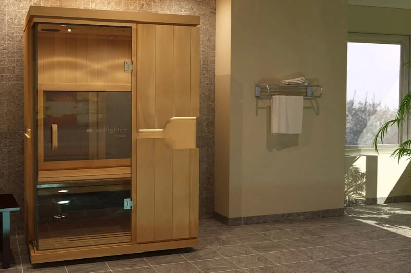 Sunlighten mPulse Believe infrared sauna installed in a modern bathroom with light wood exterior, glass door, digital control panel, and premium finishes complementing the spa-like environment with towel rack nearby.